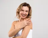 Uśmiechnięta kobieta z widocznymi zmarszczkami wokół oczu, otulona białym ręcznikiem, dotyka ramienia.