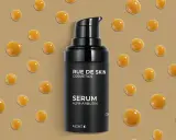 Serum z alfa-arbutyną od RUE DE SKIN. Krople serum unoszą się na beżowym tle, tworząc efekt kropli rosy.